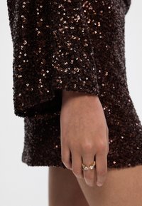 Smešasti smešasti smehani haljina s širokim rukavima, ističući ruku koja nosi zlatni prsten s dva prozirna kamena. Sjaj ističe teksturiranu površinu haljine.