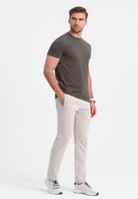 Olivengrünes T-Shirt mit kurzen Ärmeln und Tasche, kombiniert mit hellgrauen, schmal zulaufenden Hosen und weißen Mesh-Sneakern mit schwarzen Akzenten.