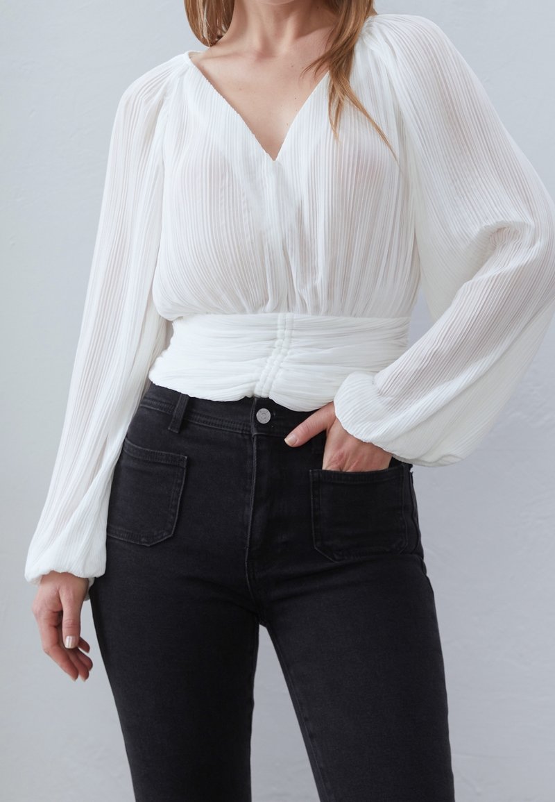 Blusa blanca con pliegues, de manga larga y escote en V profundo, combinada con jeans negros ajustados de cintura alta. Tela texturizada y detalle de cintura fruncida.