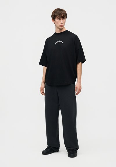 Noor mees seisab, kandes lahtisi musta värvi pükse, musta ülejate suurusega T-särki valge kirjaga ning musti kingi, vaatab veidi vasakule ühtlasel taustal.