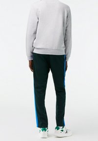Sudadera gris con puños acanalados, combinada con pantalones verde oscuro que tienen rayas laterales azules y zapatillas deportivas blancas con acentos verdes.