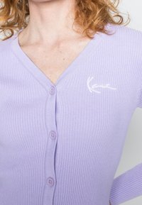 Lilaresli v-aukkoinen neuletakki, jossa on vaaleanpunaiset napit ja valkoinen kirjailtu logo rinnassa. Pehmeä pinta ja istuva malli.