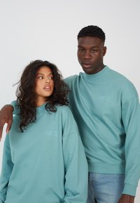 Lyse teal crewneck sweatshirt i myk stoff med brodert logo på brystet, som har en avslappet passform og ribbestrikkede mansjetter.