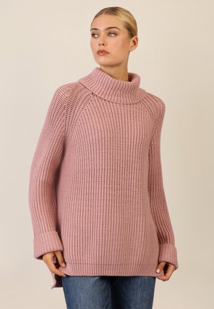 Rosa gestrickter Pullover mit einem Schalkragen, gerippter Struktur, langen Ärmeln und seitlichen Schlitzen, kombiniert mit blauen Jeans.