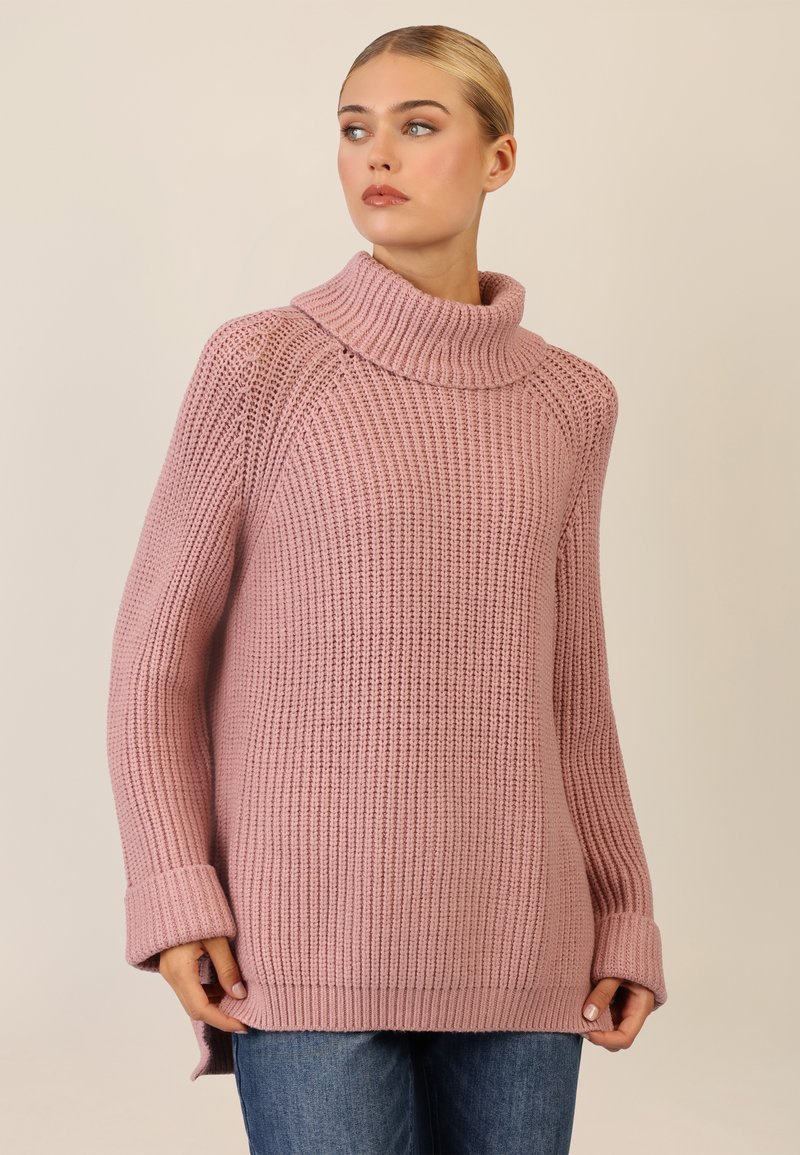 Rosa gestrickter Pullover mit einem Schalkragen, gerippter Struktur, langen Ärmeln und seitlichen Schlitzen, kombiniert mit blauen Jeans.