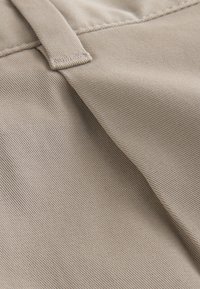 Närbild av beige tyg med sömmar och en bältesögla på ett par byxor eller shorts.