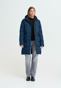 Podložena dolga plašča v temno modri barvi z kapuco, ki ima dva sprednja žepa, zadrgo na sprednji strani in teksturirano blago, oblečena čez črno pulover in sivo jeans.