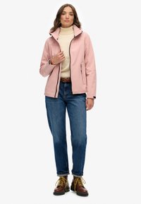 Kobieta w jasnoróżowej kurtce z zamkiem błyskawicznym na beżowym golfie, w niebieskich dżinsach z podwiniętymi nogawkami, w brązowych butach z żółtymi sznurówkami, stojąca na białym tle.