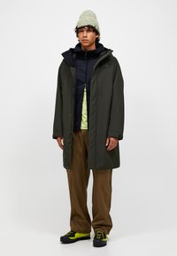 Tânăr îmbrăcat cu o pălărie verde, un palton lung de culoare verde închis, o jachetă neagră cu fermoar, pantaloni maro și ghete de drumeție negre cu galben neon.