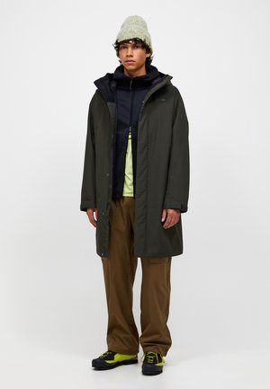 Junger Mann mit einer grünen Mütze, einem dunkelgrünen langen Mantel, einer schwarzen Zip-Jacke, braunen Hosen und schwarzen, neongelben Wanderschuhen.