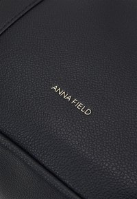 Sort tekstureret lædertaske med en glat finish, der har en fremtrædende guldfarvet "ANNA FIELD" logo. Rene linjer og moderne design.
