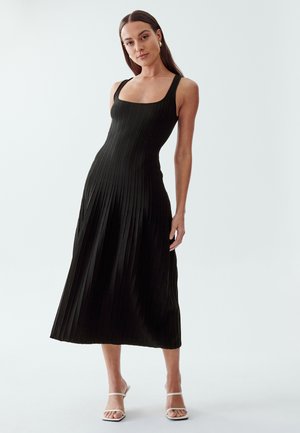 Black pleated midi dress with a square neckline and wide straps, made from a textured fabric. Model paired with white sandals.