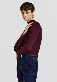 Top lungo a maniche ribbed color borgogna con collo alto; modello abbinato a jeans a vita alta blu scuro con dettagli in oro sui bottoni e sulle cuciture.