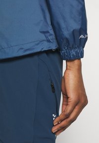 Gros plan sur la main d'une personne reposant à côté de son corps, portant une veste bleue avec un poignet élastique et un pantalon bleu avec une poche zippée.