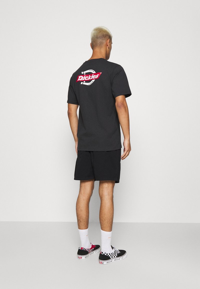 Maglietta nera a maniche corte con un grande logo rosso "Dickies" sulla schiena, indossata con pantaloni corti neri e scarpe a scacchi.