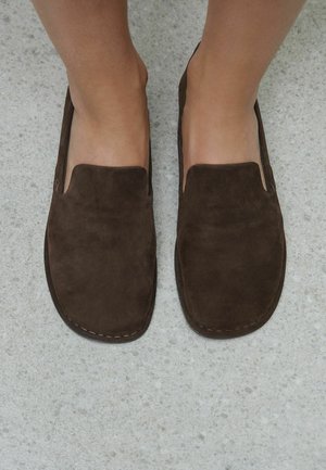 Sötétbarna velúr felcsúsztatható mokaszint viselő lábak világosszürke foltos padlón.