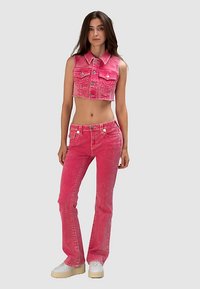 Różowa, dżinsowa krótka kamizelka z dopasowanymi, rozszerzanymi jeansami. Posiada guziki, przednie kieszenie i wyblakłą fakturę, zestawiona z białymi butami na platformie.