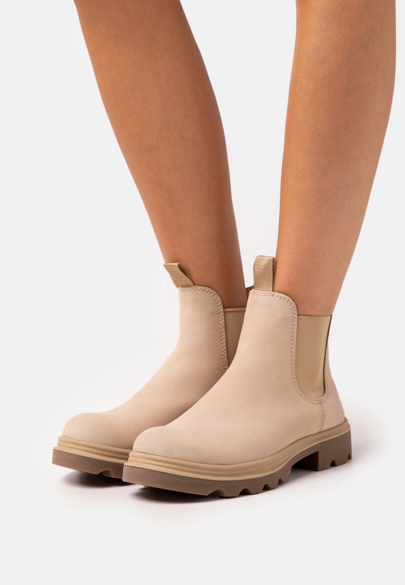 Bottes beiges à la cheville avec languettes de traction, panneaux latéraux élastiques et semelles épaisses marron, portées sur des jambes nues sur fond blanc.