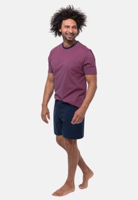 Un homme souriant avec des cheveux bouclés marche pieds nus, portant un t-shirt rayé bordeaux et un short bleu marine sur un fond blanc uni.