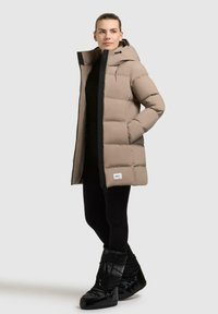 Puffer coat in light taupe with a hood, zip closure, and black accents. Features quilted design, side pockets, and a branded label at the hem.