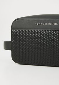 Black rectangular bag with a woven texture on the lower half and smooth leather on top. Silver "TOMMY HILFIGER" logo on the side.