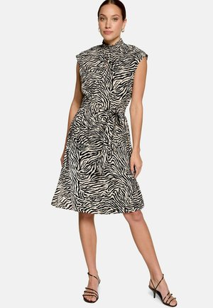 Femme portant une robe sans manches noir et beige à imprimé zèbre, avec un col montant et un lien à la taille, associée à des talons noirs à brides.