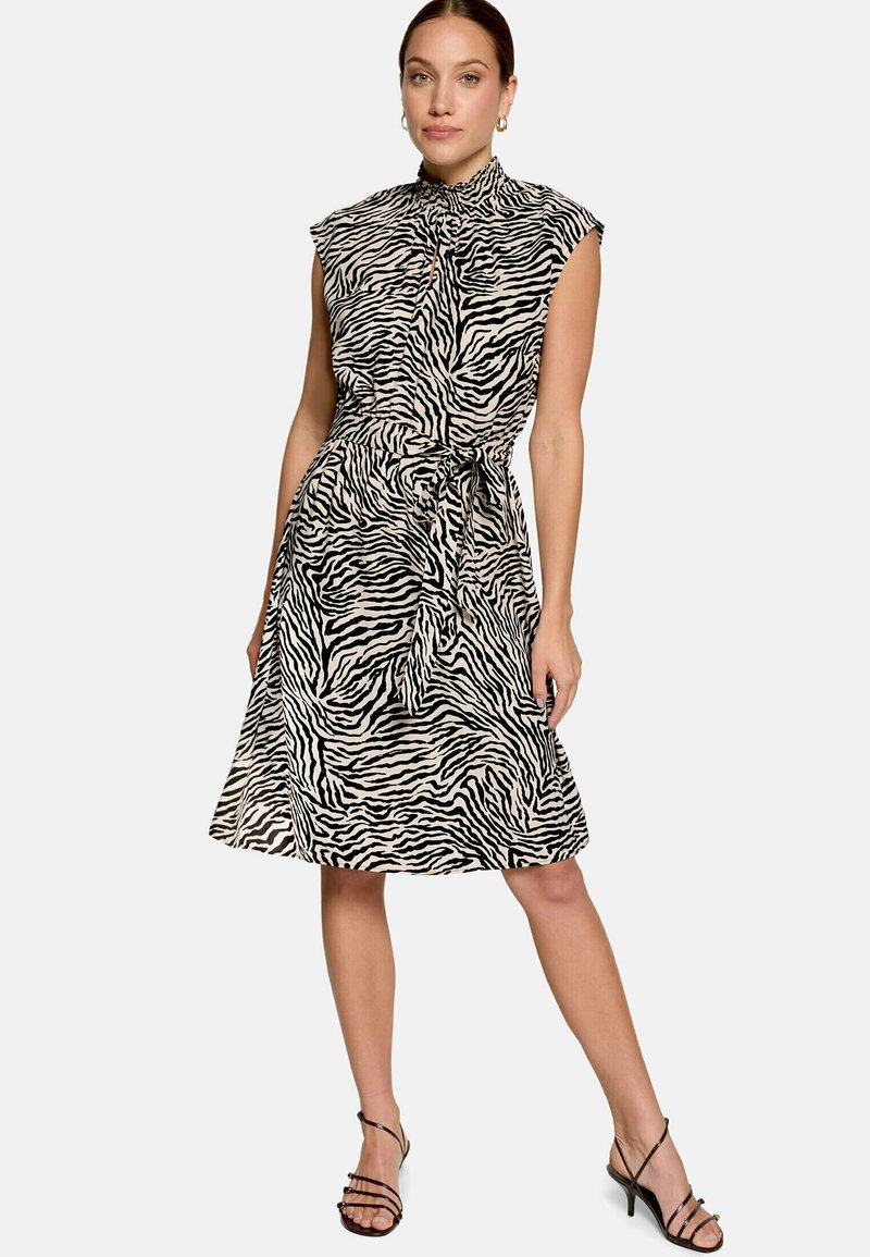 Femme portant une robe sans manches noir et beige à imprimé zèbre, avec un col montant et un lien à la taille, associée à des talons noirs à brides.