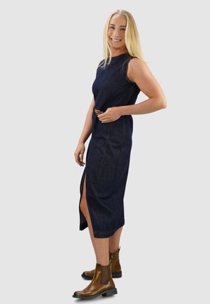 Marineblauwe denim midi-jurk met een mouwloos ontwerp, een zijsplit en een aansluitende tailleband, gecombineerd met bruine enkelbotjes.