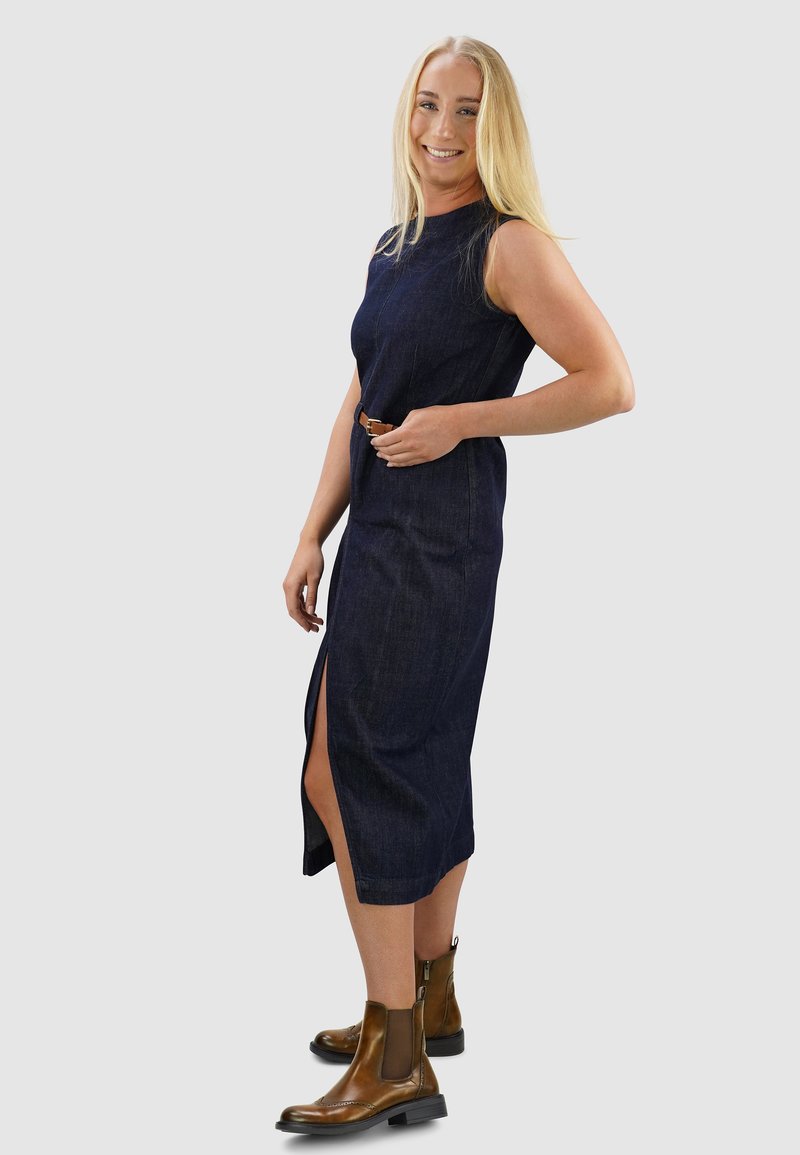 Marineblaues Denim-Midikleid mit ärmellosem Design, seitlichem Schlitz und taillierter Taille, kombiniert mit braunen Ankle Boots.