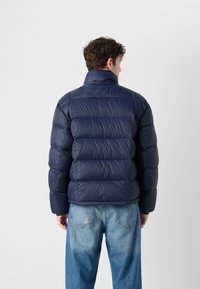 A person with curly hair wears a navy puffer jacket and light blue jeans, seen from the back against a plain white background.