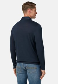 Hombre vestido con una camiseta de manga larga azul marino y jeans azules, de espaldas, frente a un fondo liso claro.
