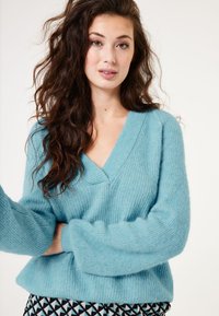 Hellblauer Strickpullover mit tiefem V-Ausschnitt, lockerer Passform und strukturiertem Stoff. Mit weiten Ärmeln und geripptem Saum. Kombiniert mit gemusterten Hosen.