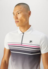 Man with short hair wearing white polo shirt with zip front and horizontal gray, pink, and black stripes, looking to the side against a plain background.