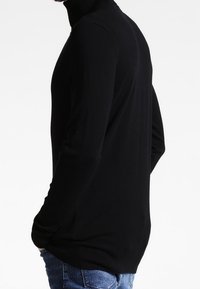 Svart polotröja med långa ärmar, tillverkad av ett slätt tyg. Har en figurnära design och ribbade muddar och fåll. Bärs med blå jeans.