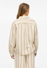 Camisa beige de gran tamaño con rayas verticales marrones, puños abotonados y cuello con solapa. Tiene un corte relajado y una textura suave.