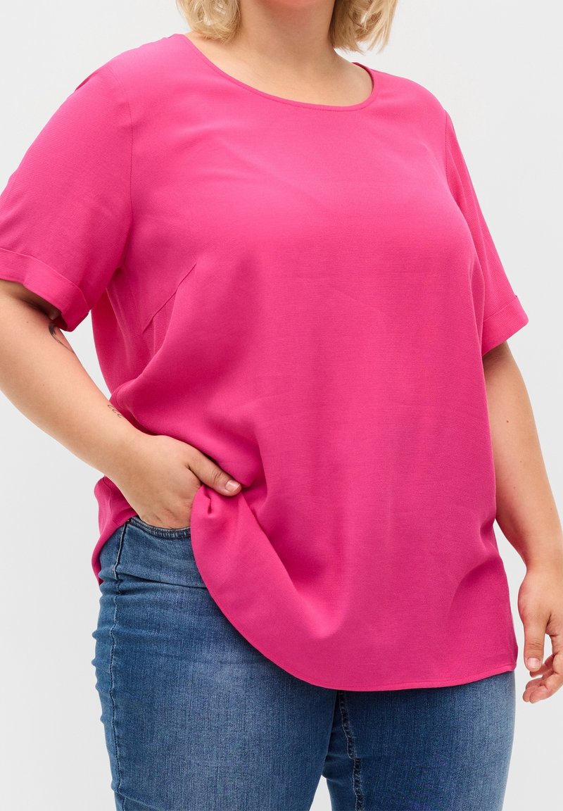 Blusa fucsia a maniche corte in tessuto leggero, caratterizzata da un colletto rotondo e fessure laterali per una vestibilità rilassata, abbinata a jeans blu.