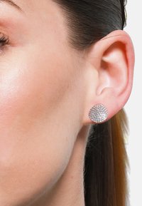 Runde silberne Steckerohrringe mit einer strukturierten Oberfläche, die mit kleinen, funkelnden Steinen bedeckt ist, vor einem Nahaufnahme eines weiblichen Ohres.