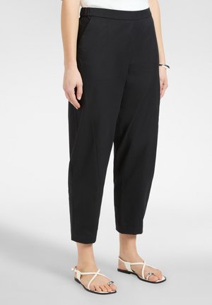 Kobieta ubrana w czarne spodnie zwężane ku dołowi, białe sandały na paski oraz biały top, stojąca na tle jednolitego jasnego tła.