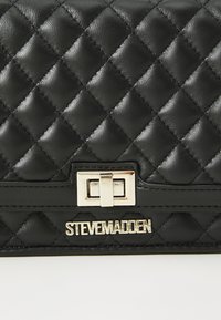 Crn quiltani umetni usnjeni denarnici z zlatim pravokotnim zapiralom in embosiranim imenom blagovne znamke "STEVE MADDEN" na dnu.