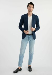 Granatowa marynarka z klapami w kształcie notcha, biała koszulka, jasnoniebieskie dżinsy i czarne mokasyny. Marynarka ma jedną kieszeń na piersi i dopasowany krój.