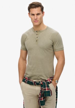 Olivenfarvet kortærmet henley t-shirt med tre sorte knapper, parret med oprullede kaki bukser og en rød-grønternet skjorte bundet om taljen.