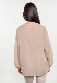 Blusa beige chiaro con maniche lunghe, tessuto a trama morbida, scollatura rotonda e polsini arricciati. Le cuciture visibili creano una siluetta rilassata.