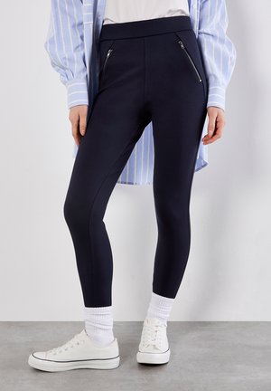 Leggings navy a vita alta, con due tasche con zip sul davanti, abbinati a una camicia a righe azzurra chiara e sneakers bianche.