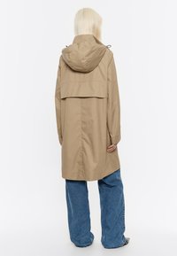 Beige regenjas met een capuchon, voorzien van een rugpand en lange mouwen. Gecombineerd met losse blauwe denim jeans, met minimale details.