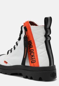 Botte blanche montante avec lacets noirs, semelle épaisse noire, fermeture éclair orange étiquetée "UNLOCKED," et languette noire avec le texte "FREEDOM".