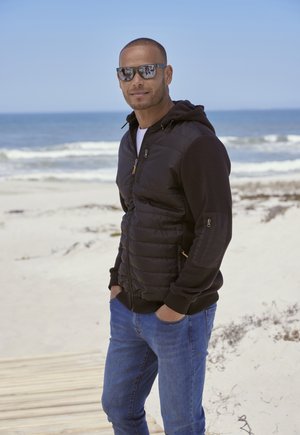 Giacca a vento nera con cappuccio, dettagli con zip e polsini elasticizzati, abbinata a jeans in denim blu; sullo sfondo si vede una spiaggia.