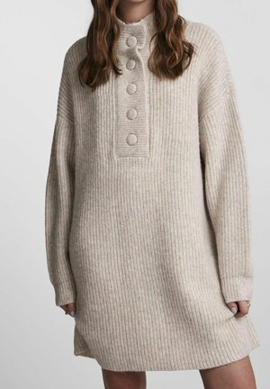 Femme portant une robe-pull en maille beige à manches longues avec cinq grands boutons recouverts sur la patte de boutonnage devant.