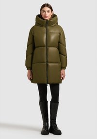 Olijfgroene donsjas met een hoge kraag, verticale vlakken en een ritssluiting aan de voorkant. Gecombineerd met zwarte legging en kniehoge laarzen.