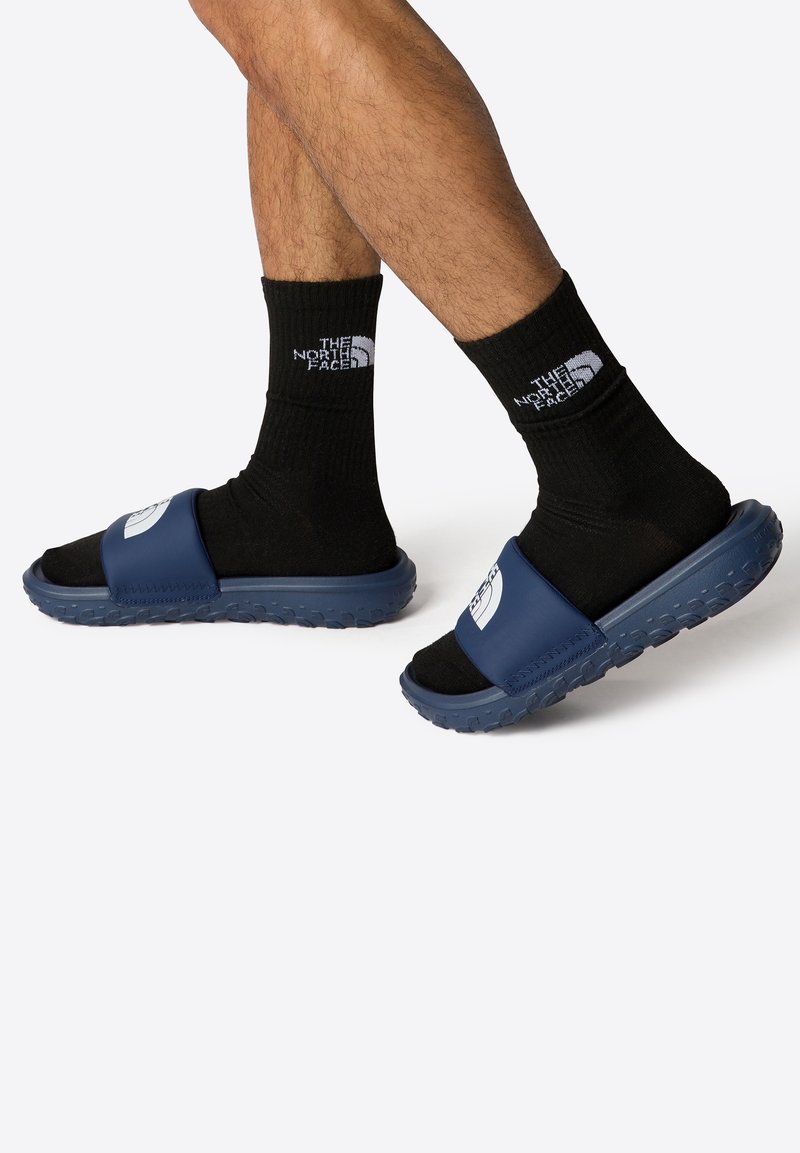 Chanclas azules con una tira ancha y una suela texturizada, combinadas con calcetines negros que tienen un logo blanco en el puño.