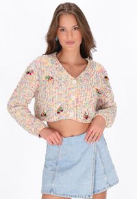 Junge Frau in einer bunten, gestrickten, kurzen Cardigan mit Pompons und einem hellblauen Jeans-Minikleid, die mit den Händen auf den Hüften posiert.